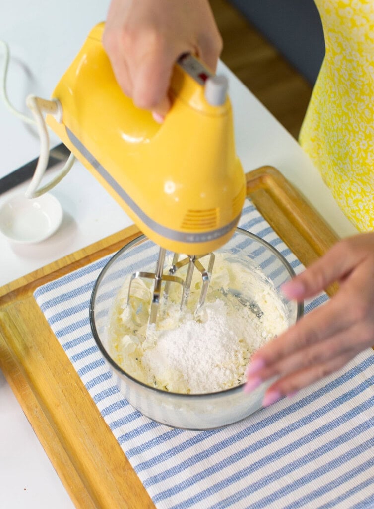 A person uses a yellow hand mixer to blend ingredients, including flour, in a glass bowl on a blue-striped cloth and wooden tray.