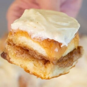 A hand holding a piece of cinnamon roll topped with creamy white icing, showing the swirled layers of cinnamon and sugar inside.
