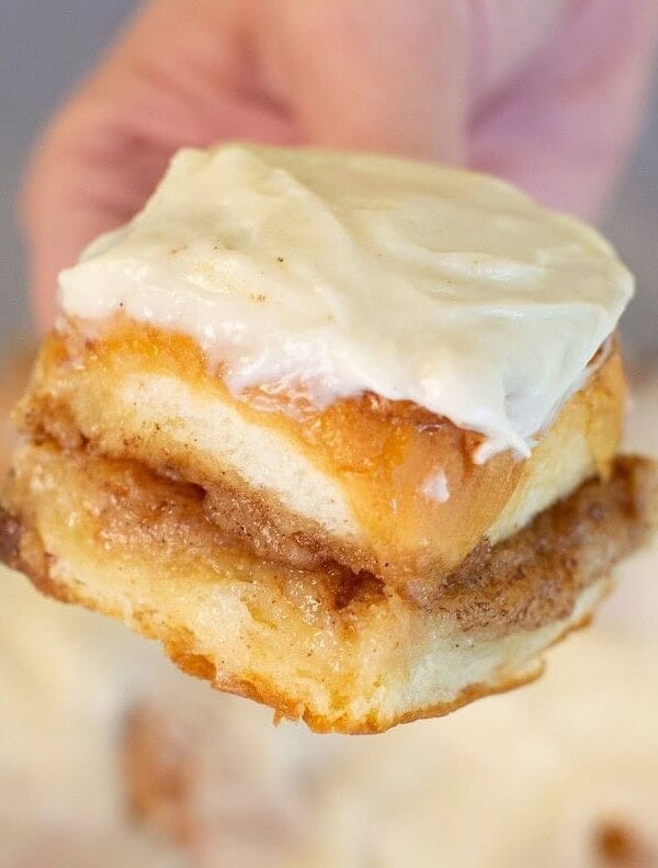 A hand holding a piece of cinnamon roll topped with creamy white icing, showing the swirled layers of cinnamon and sugar inside.