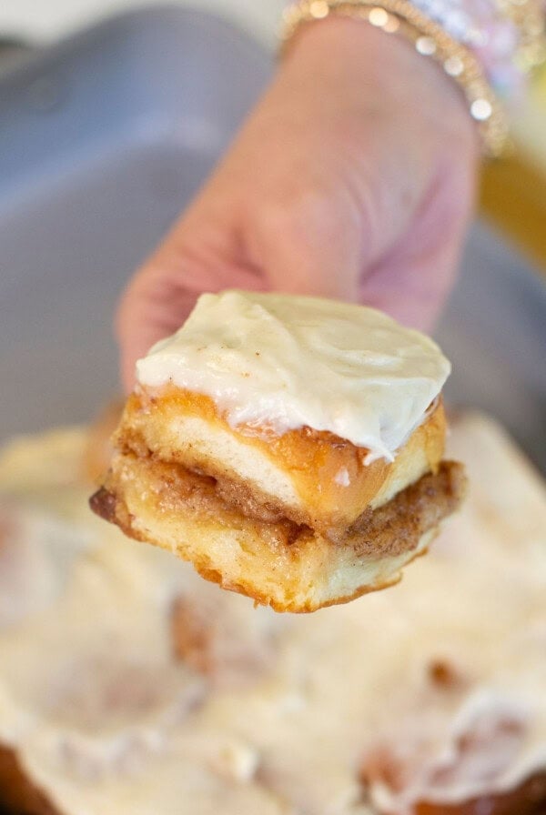 A hand holding a frosted cinnamon roll piece with creamy icing, with more cinnamon rolls in a baking pan in the background.