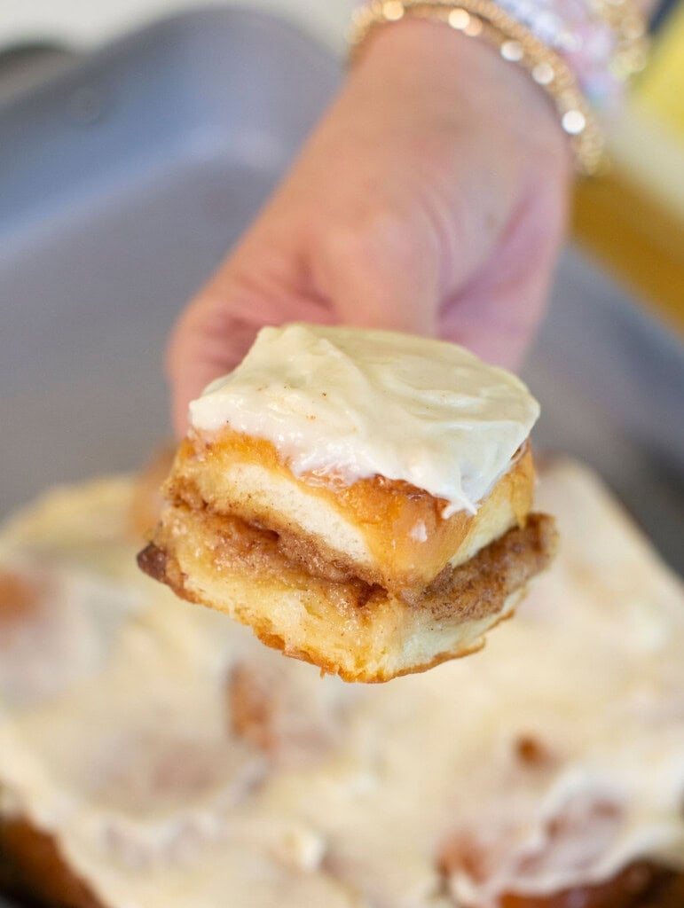A hand holding a frosted cinnamon roll piece with creamy icing, with more cinnamon rolls in a baking pan in the background.