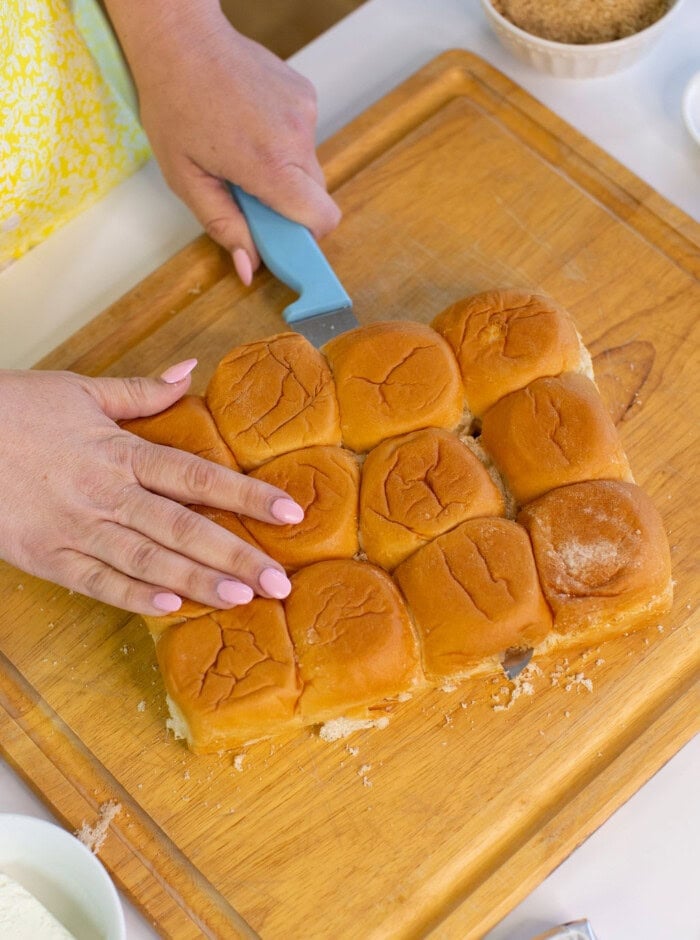 A person with light pink nail polish slices a sheet of soft dinner rolls on a wooden cutting board using a blue-handled knife. Bowls with ingredients are visible nearby.