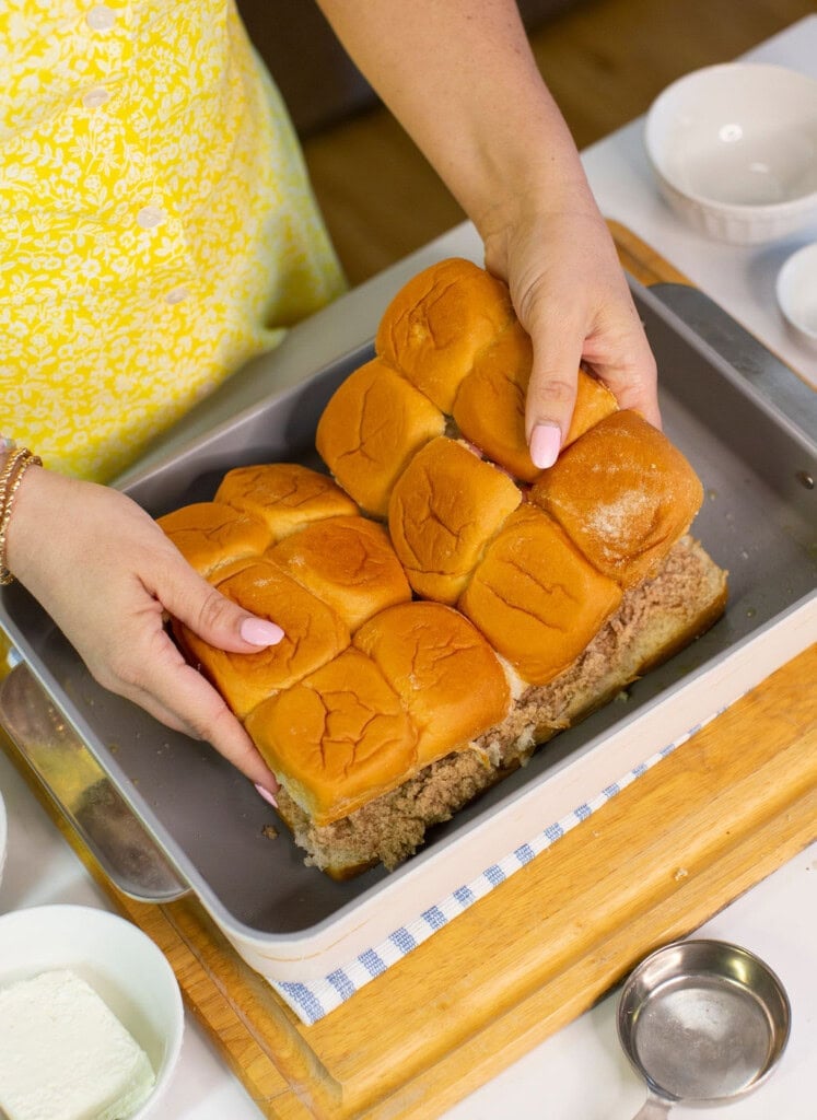 A person in a yellow patterned apron places a layer of connected slider buns over shredded meat in a baking pan, surrounded by small bowls on a kitchen counter.