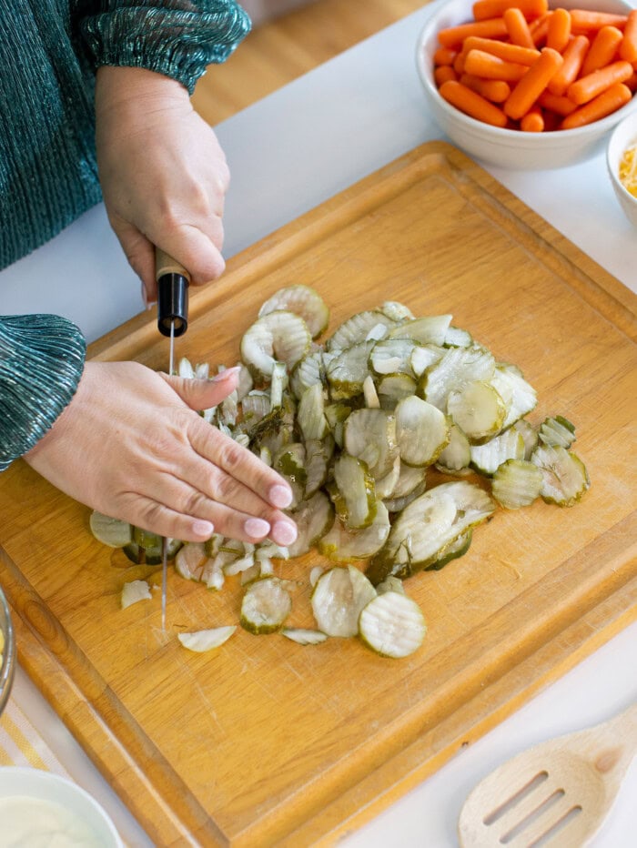 A person slices pickles on a wooden cutting board with a knife. Nearby are bowls containing baby carrots and other ingredients.