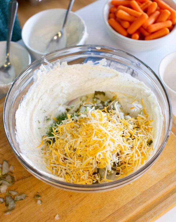 A glass bowl filled with a creamy dip mixture, topped with shredded cheese, chopped herbs, and pickles sits on a wooden cutting board. In the background, a bowl of baby carrots and two empty bowls with spoons are visible.
