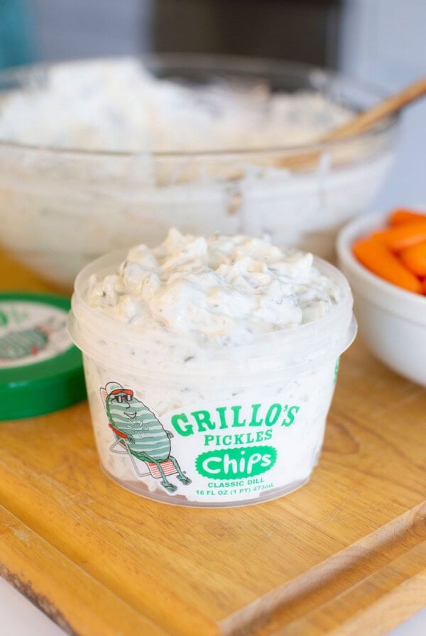 A container of Grillo’s Pickles Classic Dill Chips dip sits on a wooden board, with chopped carrots in a white bowl and a mixing bowl with a wooden spoon in the background.