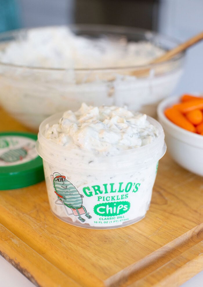 A container of Grillo’s Pickles Classic Dill Chips dip sits on a wooden board, with chopped carrots in a white bowl and a mixing bowl with a wooden spoon in the background.