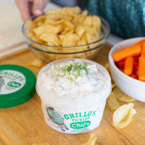 A container of Grillo’s Pickles chips dip with dill on top sits on a wooden surface, surrounded by potato chips, a bowl of baby carrots, and a bowl of chips. A person’s hand reaches toward the chip bowl.