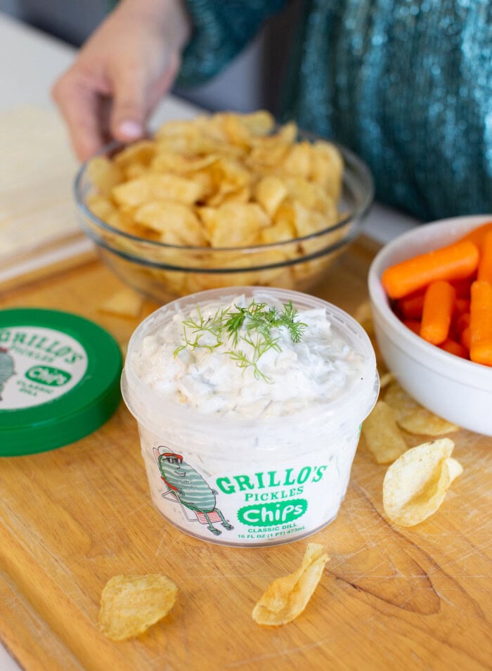 A container of Grillo’s Pickles chips dip with dill on top sits on a wooden surface, surrounded by potato chips, a bowl of baby carrots, and a bowl of chips. A person’s hand reaches toward the chip bowl.