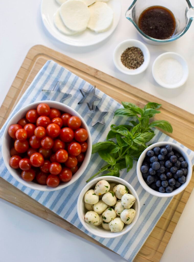 A cutting board with bowls of cherry tomatoes, blueberries, mozzarella balls, fresh basil, and small containers of spices and balsamic dressing, set on a blue-striped cloth. Slices of mozzarella are also visible.