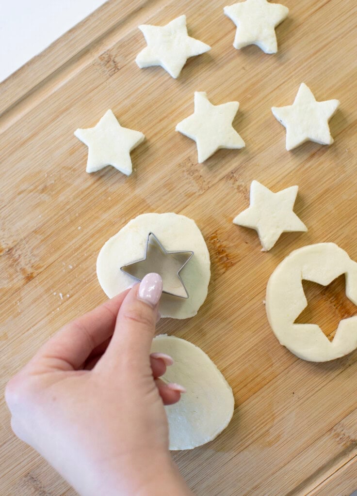 A hand uses a star-shaped cookie cutter to cut star shapes from dough on a wooden board. Several star-shaped dough pieces are arranged on the board.