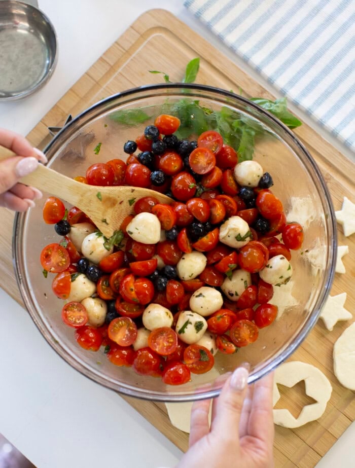A person mixes a salad of cherry tomatoes, mozzarella balls, blueberries, and herbs in a glass bowl with a wooden spoon on a kitchen counter beside a cutting board with star-shaped dough.