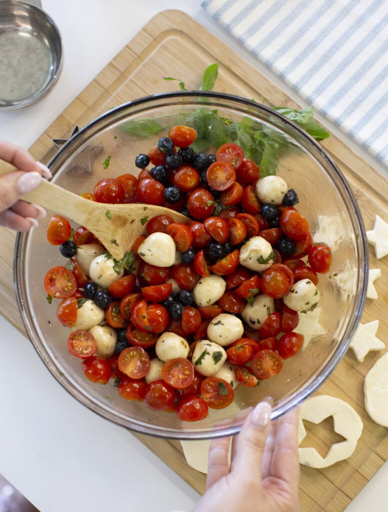 A person mixes a salad of cherry tomatoes, mozzarella balls, blueberries, and herbs in a glass bowl with a wooden spoon on a kitchen counter beside a cutting board with star-shaped dough.