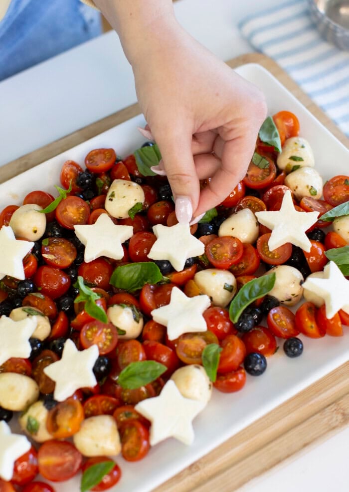 A hand places a star-shaped piece of cheese on a platter of cherry tomatoes, mozzarella balls, blueberries, and fresh basil. The colorful salad is arranged on a white rectangular dish.