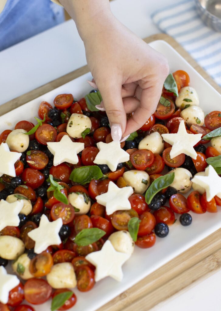 A hand places a star-shaped piece of cheese on a platter of cherry tomatoes, mozzarella balls, blueberries, and fresh basil. The colorful salad is arranged on a white rectangular dish.