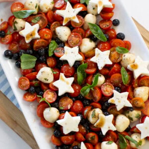 A rectangular platter with cherry tomatoes, blueberries, mozzarella balls, fresh basil, and star-shaped mozzarella slices, drizzled with dressing. A hand is holding the edge of the plate.