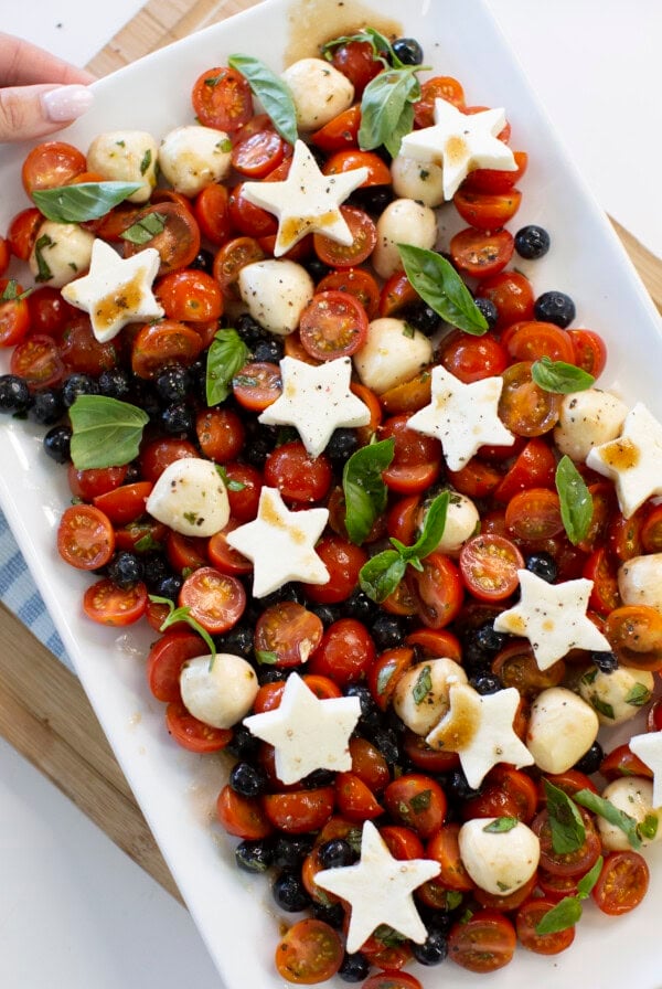 A rectangular platter with cherry tomatoes, blueberries, mozzarella balls, fresh basil, and star-shaped mozzarella slices, drizzled with dressing. A hand is holding the edge of the plate.