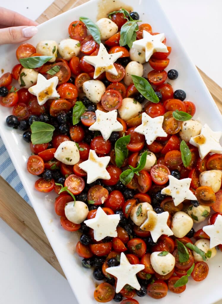 A rectangular platter with cherry tomatoes, blueberries, mozzarella balls, fresh basil, and star-shaped mozzarella slices, drizzled with dressing. A hand is holding the edge of the plate.