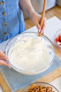 A person in a blue buttoned dress is mixing whipped cream in a glass bowl with a spatula. Nearby, there are bowls of sliced strawberries and pretzels on a light blue placemat.