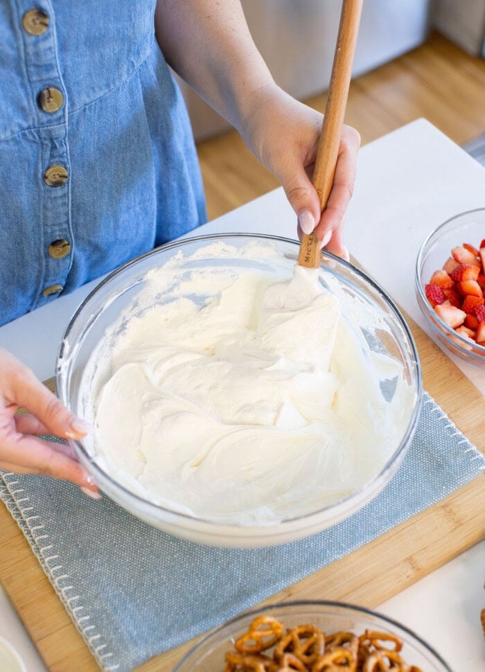 A person in a blue buttoned dress is mixing whipped cream in a glass bowl with a spatula. Nearby, there are bowls of sliced strawberries and pretzels on a light blue placemat.