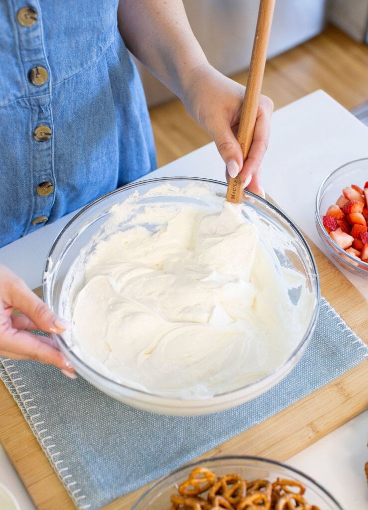 A person in a blue buttoned dress is mixing whipped cream in a glass bowl with a spatula. Nearby, there are bowls of sliced strawberries and pretzels on a light blue placemat.