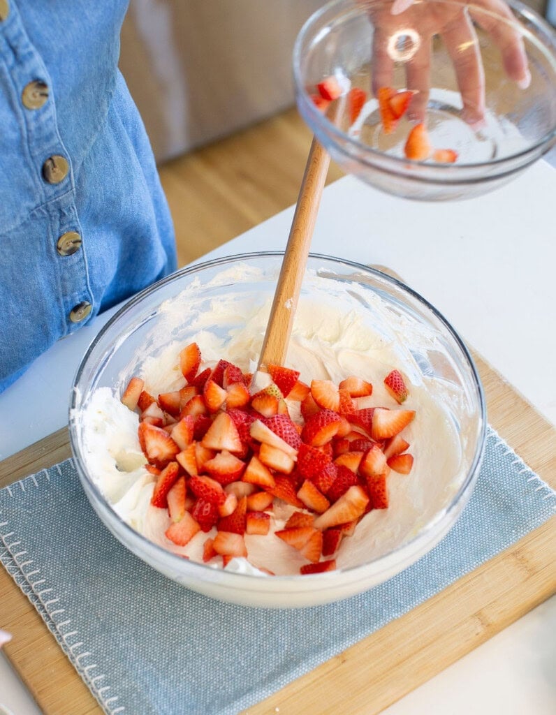 A person pours a bowl of chopped strawberries into a larger bowl filled with creamy batter, stirring with a wooden spoon, on a wooden board with a blue placemat.