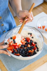 A person in a blue buttoned dress stirs a glass bowl of whipped cream, blueberries, and chopped strawberries on a wooden board, preparing a fruit dessert.