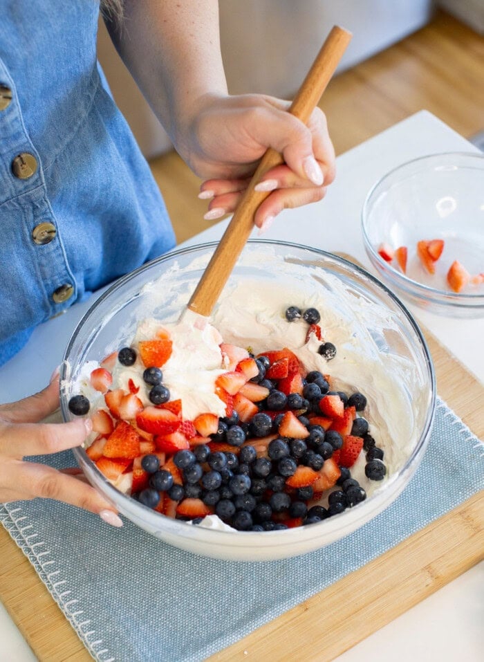 A person in a blue buttoned dress stirs a glass bowl of whipped cream, blueberries, and chopped strawberries on a wooden board, preparing a fruit dessert.