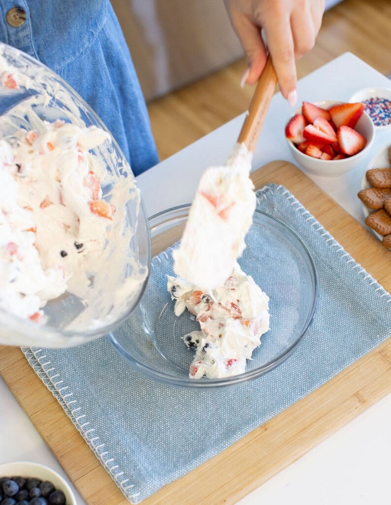 A person uses a spatula to transfer a creamy fruit mixture with berries from a bowl into a glass dish. Other ingredients like strawberries, sprinkles, and cookies are on the table nearby.