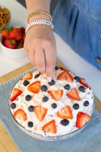 A person’s hand is topping a bowl of creamy dessert with sliced strawberries and blueberries. The bowl sits on a wooden board with a blue cloth, and a bowl of strawberries and pretzels are in the background.