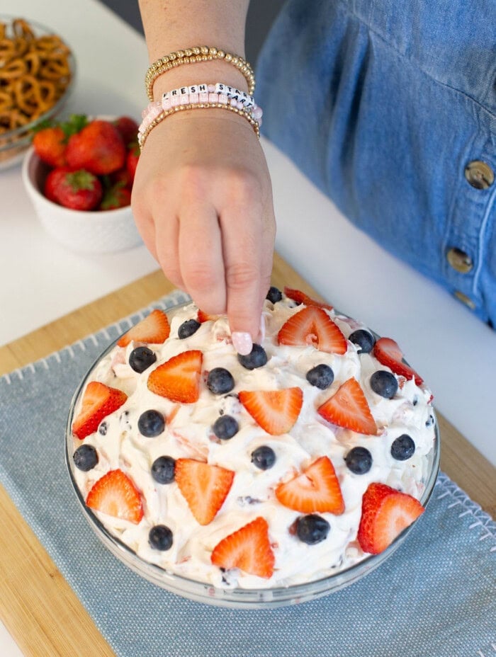 A person’s hand is topping a bowl of creamy dessert with sliced strawberries and blueberries. The bowl sits on a wooden board with a blue cloth, and a bowl of strawberries and pretzels are in the background.
