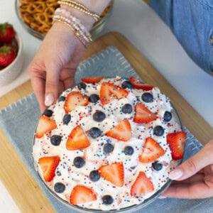 A person holding a dessert topped with whipped cream, sliced strawberries, blueberries, and red, white, and blue sprinkles, on a blue cloth. Pretzels and strawberries are in bowls nearby.