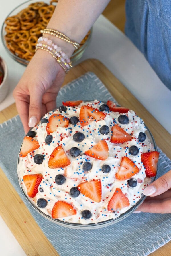 A person holding a dessert topped with whipped cream, sliced strawberries, blueberries, and red, white, and blue sprinkles, on a blue cloth. Pretzels and strawberries are in bowls nearby.