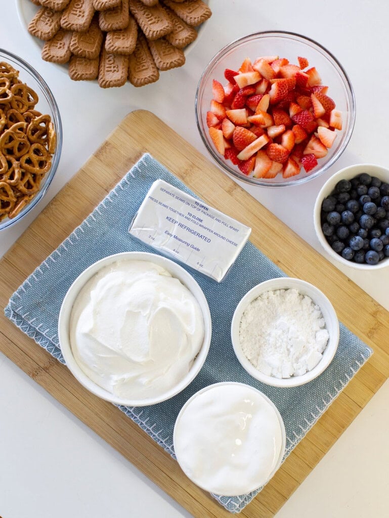 A wooden board with bowls of whipped cream, powdered sugar, and cream cheese on a blue cloth. Nearby are dishes of sliced strawberries, blueberries, pretzels, and rectangular cookies.