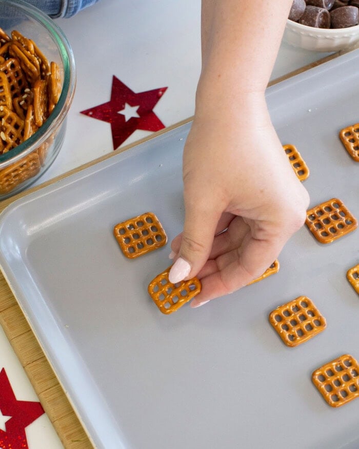 A hand places square pretzels on a baking sheet, with more pretzels in a glass bowl nearby and decorative red star cutouts on the table.