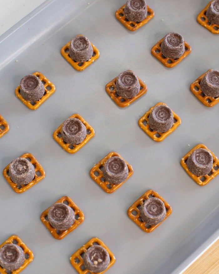 A baking sheet lined with square pretzels, each topped with a chocolate-covered caramel candy, arranged in neat rows before baking.