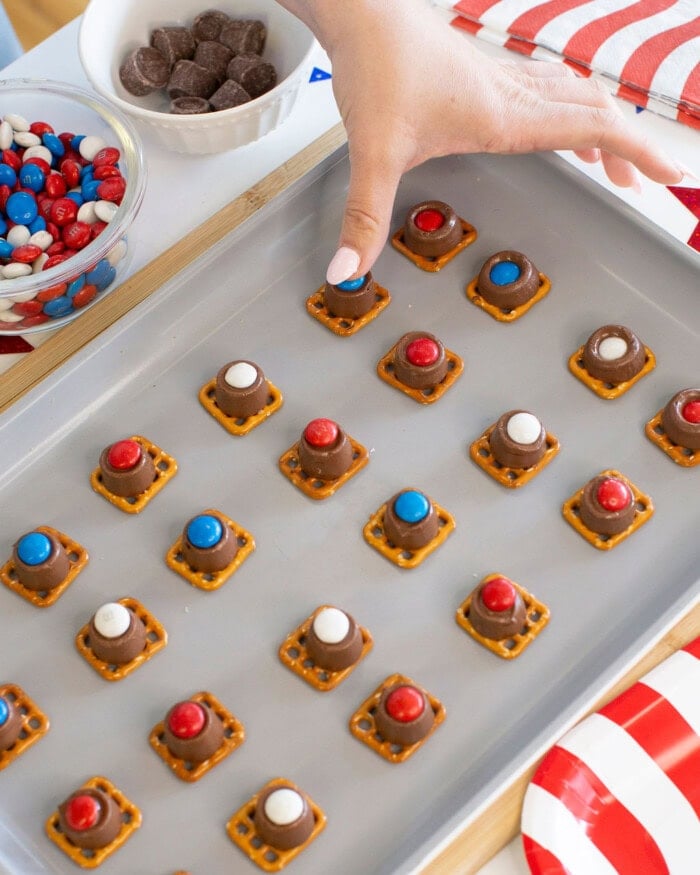 A hand places a red candy-topped chocolate on a small pretzel square on a baking sheet with similar treats; bowls of candies and chocolate pieces are nearby, along with a red and white striped napkin.