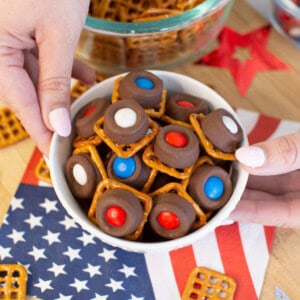 A bowl of pretzel bites topped with chocolate and red, white, and blue candies is held above an American flag napkin, with more pretzels and festive decorations in the background.