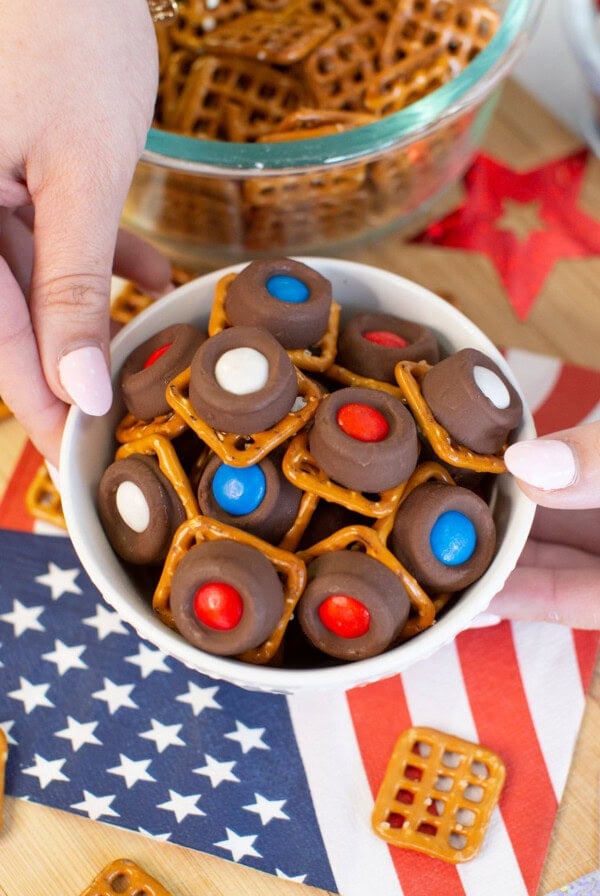 A bowl of pretzel bites topped with chocolate and red, white, and blue candies is held above an American flag napkin, with more pretzels and festive decorations in the background.