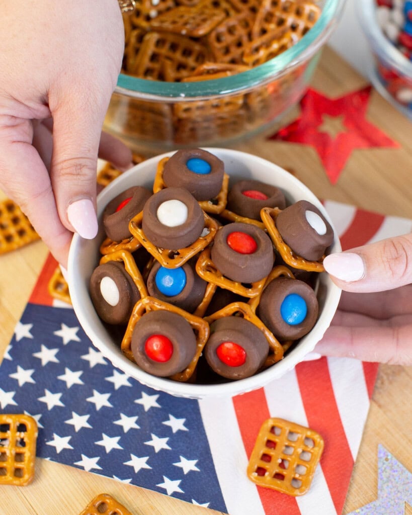 A bowl of pretzel bites topped with chocolate and red, white, and blue candies is held above an American flag napkin, with more pretzels and festive decorations in the background.