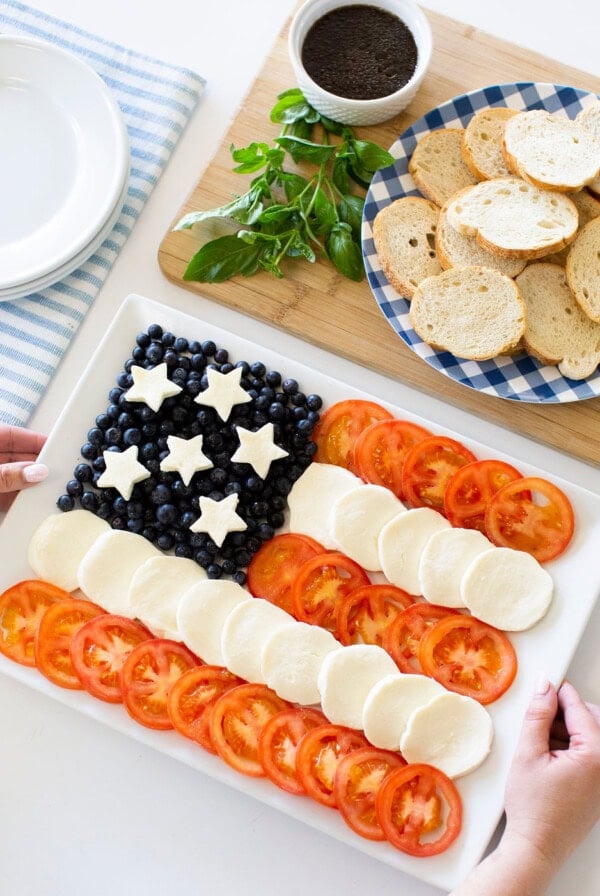 A rectangular platter displays an American flag made of blueberries with mozzarella stars, and alternating rows of mozzarella and tomato slices; nearby are basil, a bowl of dip, and a plate of sliced baguette.