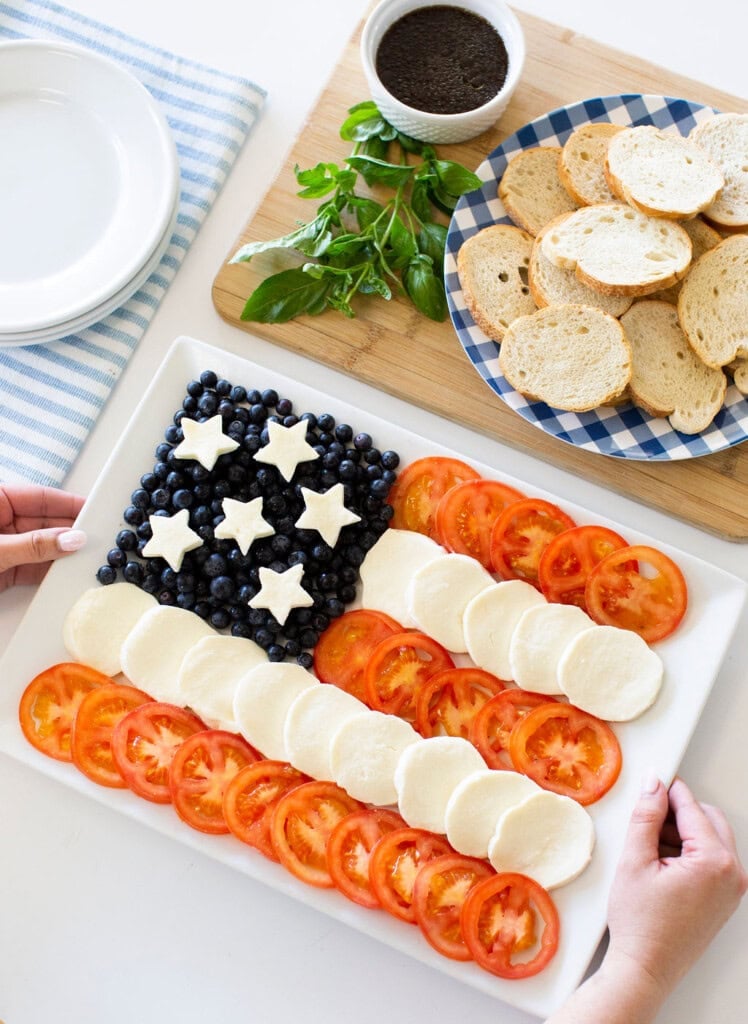 A rectangular platter displays an American flag made of blueberries with mozzarella stars, and alternating rows of mozzarella and tomato slices; nearby are basil, a bowl of dip, and a plate of sliced baguette.