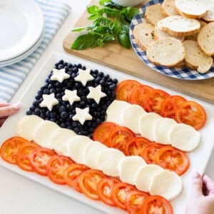 A platter arranged like the U.S. flag, with blueberries and star-shaped cheese for the stars, and rows of sliced tomatoes and mozzarella for the stripes. Nearby are toasted bread slices and a bowl of dip.