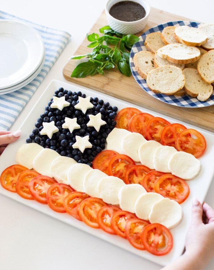A platter arranged like the U.S. flag, with blueberries and star-shaped cheese for the stars, and rows of sliced tomatoes and mozzarella for the stripes. Nearby are toasted bread slices and a bowl of dip.