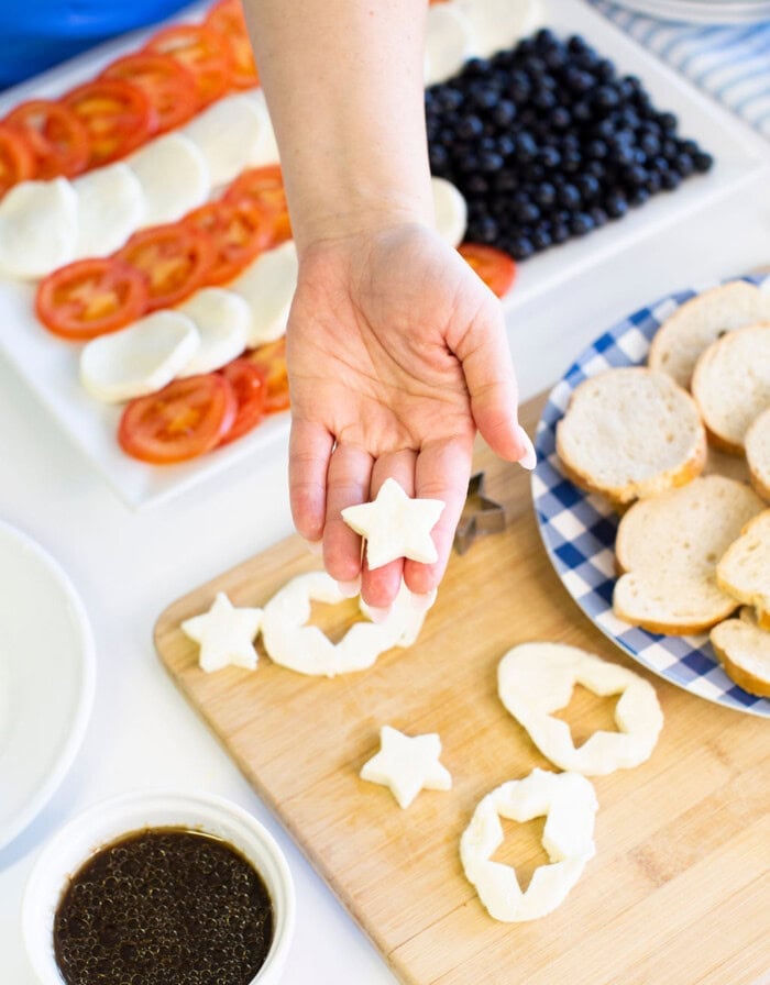 A hand holds a star-shaped mozzarella cheese slice over a cutting board with more cheese stars, bread slices, and a bowl of balsamic dressing. Sliced tomatoes, mozzarella, and blueberries are in the background.