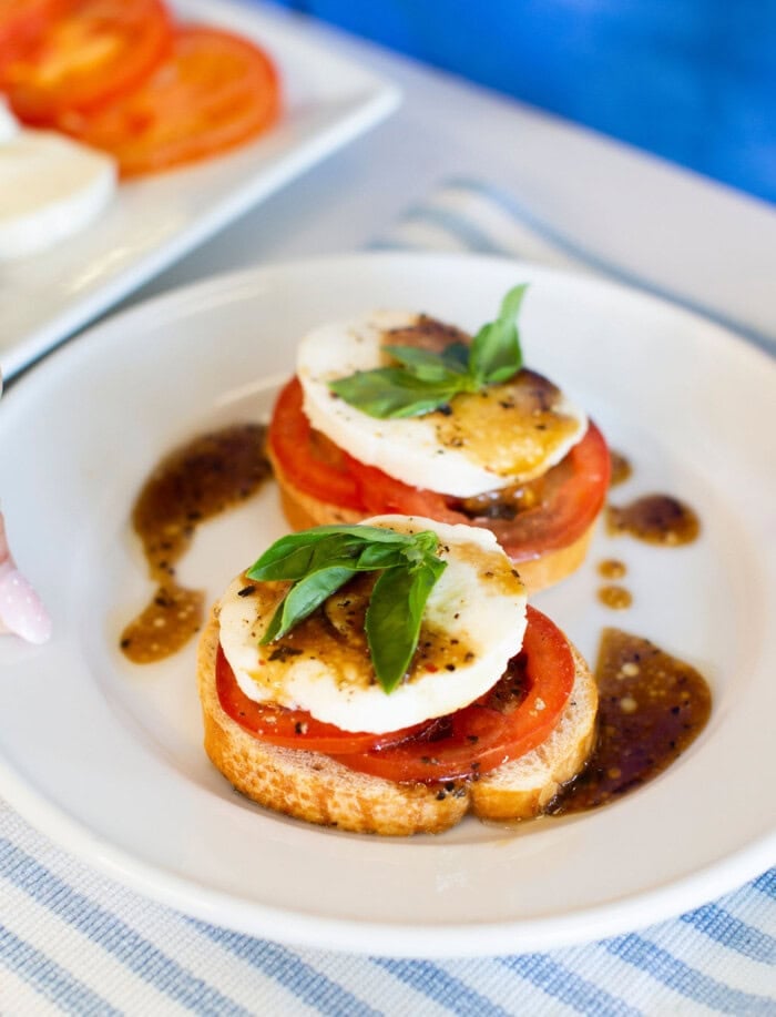 Two pieces of toasted bread topped with tomato slices, fresh mozzarella, basil leaves, and drizzled with balsamic glaze, served on a white plate with a side of extra sauce.