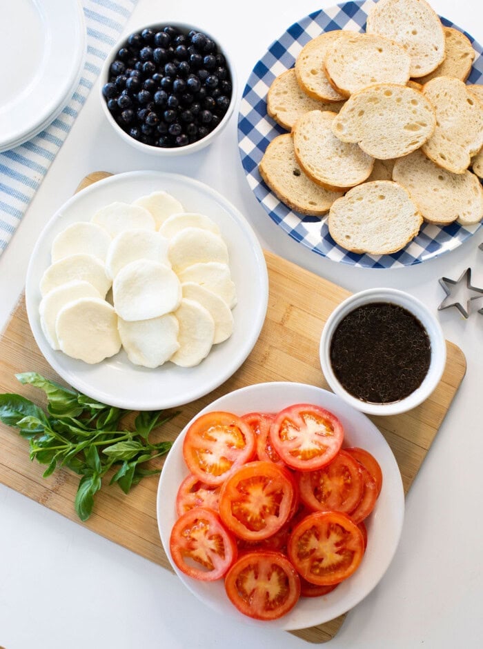 A tabletop with sliced bread, mozzarella, tomatoes, fresh basil, a bowl of blueberries, and a bowl of ground black pepper on various plates and a cutting board.