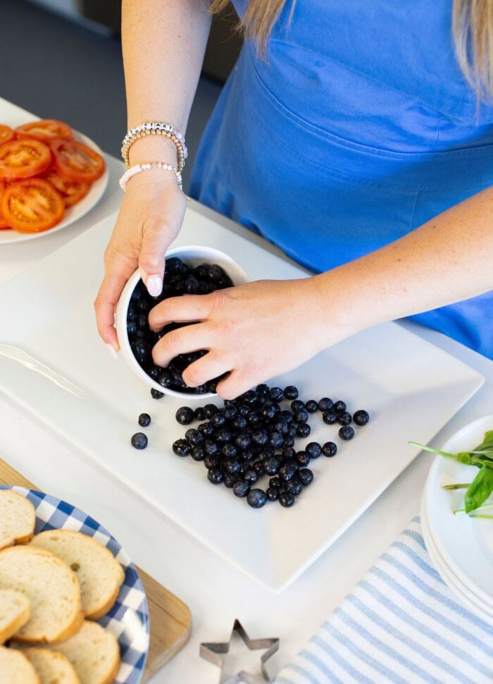 A person in a blue dress pours fresh blueberries from a bowl onto a white platter. Sliced tomatoes, bread, and leafy greens are arranged nearby on the table.