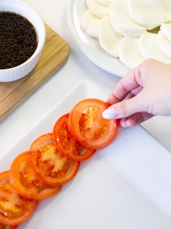A hand arranges fresh tomato slices on a white plate. Nearby, there is a bowl of dark sauce and a plate with round pieces of white bread or steamed buns.