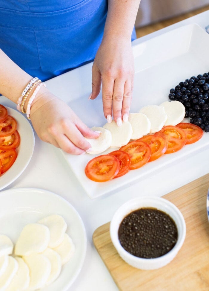 A person arranges slices of fresh mozzarella and tomato on a white platter, next to a pile of blueberries. A bowl of dark dressing and more mozzarella slices are nearby on the table.